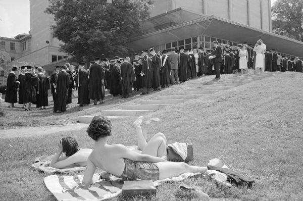 A group of 447 University of Wisconsin honor students stand in line to enter the University Memorial Union theater for the 21st annual honor's convocation ceremony. Two young women are sunbathing in the foreground on the lake shore and are watching the procession.     