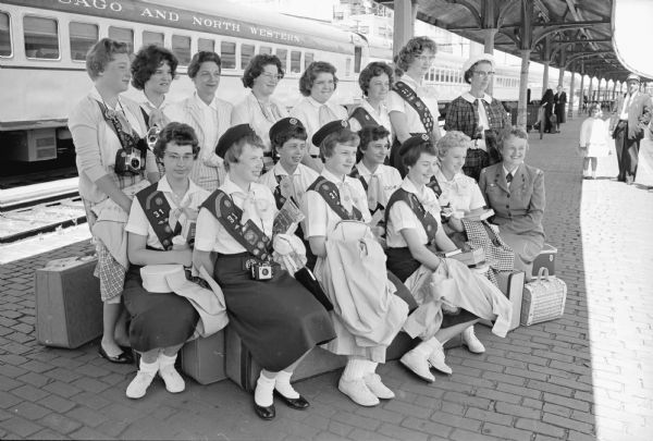 Thirteen Girl Scouts from Troop 31 from Sherman School prepare to depart on a bus to Minneapolis. The Girl Scouts will earn points for their travel badge. The troop leader and two chaperones accompany the scouts. Shown (L-R), top row, are: Brenda McGillivray, Leah Sachtjen, Mrs. R. Dolgner, chaperone, Chris Herbst, Karen Lee, Bev Tully, Elaine Larson, and Laura Conrad, chaperone; in the bottom row are: Sharon O'Kane, Judy Willis, Joan Droster, Carole Shinstine, Kathy Carmody, Rae Conrad,and Mrs. Edwin Conrad, troop leader.