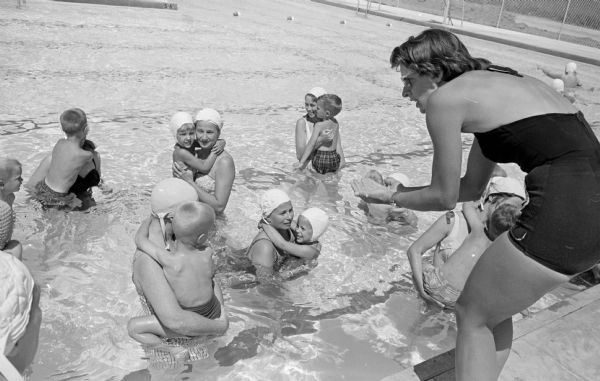 At right, Berthaida Fairbanks, physical education and Red Cross instructor, is standing on the side of a swimming pool training the women in the pool how to help their children learn to swim.