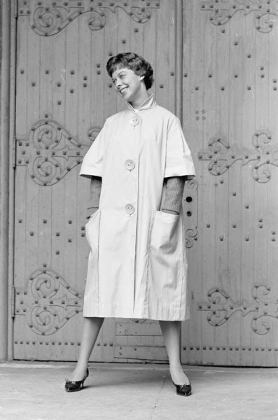 Judy Dornfeld wears a "beatnik" raincoat that doubles as a topcoat. Miss Dornfeld will be a junior at the University of Wisconsin. She is standing in front of a large door.