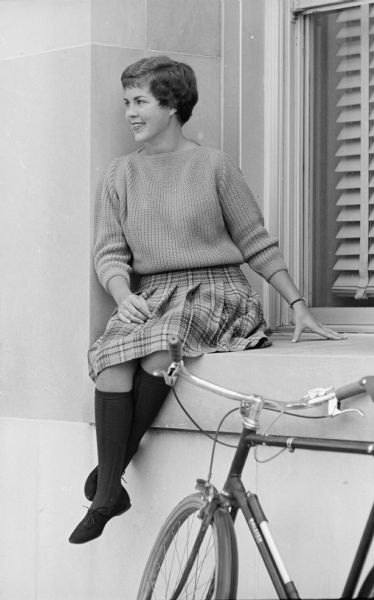 A model posing sitting on a deep window sill near a bicycle. Sara Volz teams a blue and gold plaid skirt with a gold bulky knit orlon sweater, navy blue knee socks, and suede shoes. She will be a junior at Lawrence College, in Appleton, Wisconsin.