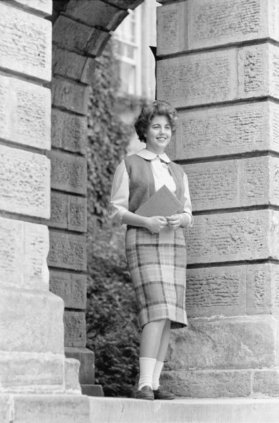Janice Clark, a freshman at the University of Wisconsin and a graduate of East high school in Madison, is shown wearing a straight stone green and gray plaid wool skirt with a matching sweater vest and a cotton blouse in a lighter tone of stone green.