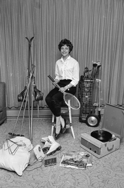 Bonnie Isabella, a freshman at the University of Wisconsin and a graduate of Madison West high school, wearing black cotton knit capri pants and a white "sissy" blouse, plus flat-heeled shoes - attire for dormitory wear and climbing Bascom hill. She also is shown with recommended accessories: skis, golf clubs, ice skates, tennis racket, radio, clock, record player and records, coat hangers, and a laundry bag.