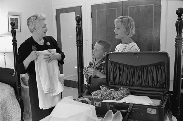 Florence Sullivan is shown packing a suitcase in her bedroom as John and Jane Sullivan, two of her grandchildren look on. Florence Sullivan, nearly 78 years old, "delighted in taking her many grandchildren (in groups of three or four at a time) on trips to all parts of the country." Their father is Arthur G. Sullivan Jr., Horicon.