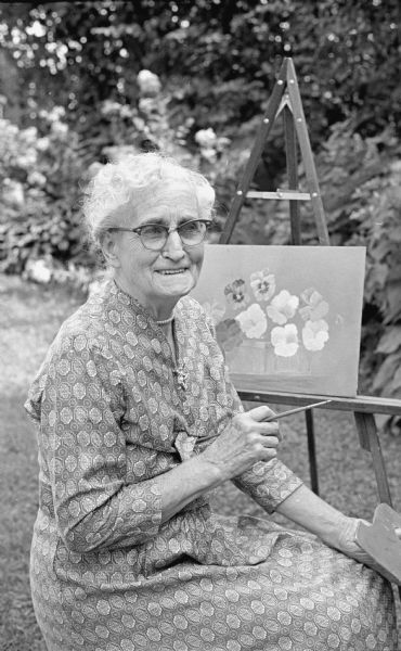 Katharine A. Church is shown seated outside in her backyard, paintbrush in hand, in front of an easel with a painting depicting flowers. A flower garden is in the background. She manages the building in which she lives, at 2107 University Avenue. She is an 89-year-old great-grandmother.