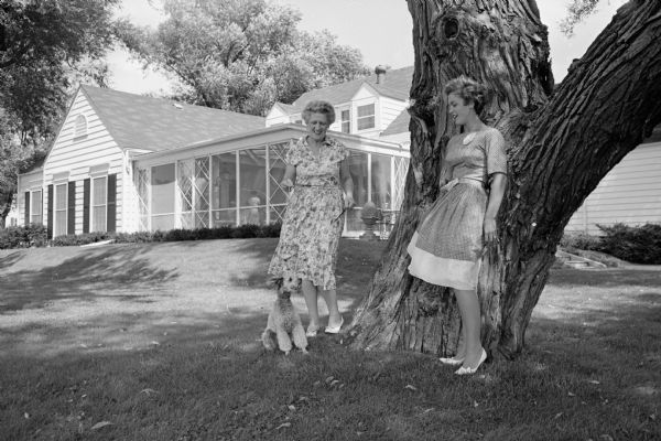 Mary Johnson (Mrs. H. Stanley Johnson), left, hostess for the Attic Angel Association benefit antique and artificial flower sale, is shown with her new daughter-in-law Marilyn Johnson (Mrs. Stanley Allen Johnson) on the spacious lawn of the elder Mrs. Johnson's home, 17 Cambridge Court, Maple Bluff, where the sale will be held. The dog is a French silver-gray poodle, Monsieur X.