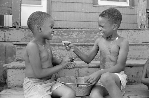 Two African-American boys shooting water pistols at each other on a hot summer day while sitting on front porch steps.