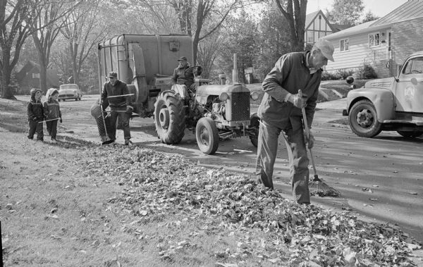 A Madison street department crew cleans leaves from the gutter along Mandan Crescent using a leaf vacuum machine as neighborhood children watch. Workers are, left to right, Carroll Gray, John Romano, and William Skolaski.
