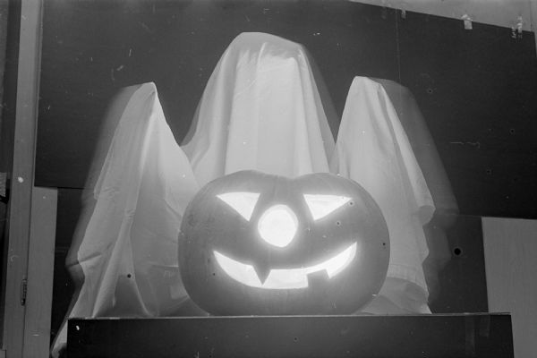 A lighted smiling carved Jack-O-Lantern with a ghost figure behind.
