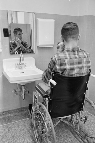 A scene at the Wisconsin Neurological Foundation, 1954 East Washington Avenue, which is observing it's 10th anniversary. Patient Jim Markl, Watertown, WI, is shown in a wheelchair shaving himself with the aid of a special hand clamp. Afflicted by a spinal injury, he has been a patient for 10 months and has re-learned several skills.