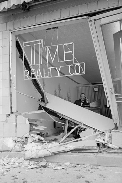 The smashed-in front facade of the Time Realty Co. office, 2434 University Avenue, as a result of a car backing up with a stuck accelerator. Seen inside the building is Lew Greisen, a company salesman.