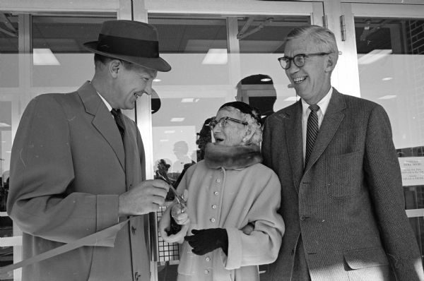 Jessie E. Manchester, widow of merchant Harry S. Manchester, prepares to snip the ribbon officially opening the third store at the Westgate Shopping Center. Looking on are Mayor Ivan Nestingen (left) and Morgan Manchester, president of the firm.
