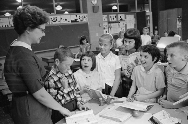 Third grade students gather around teacher Diane Schmitt's desk before school begins. Students are Jerry Tennant, Cathy Kvalheim, Vicky Seltzner, Suzanne Nickel, and Gail Kump. The boy on the far right was cropped out of the image printed in the newspaper and therefore is not identified.