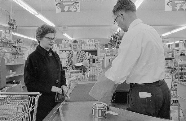 New Sun Prairie third grade teacher Diane Schmitt shops for groceries on her way home from school. The cashier is Carl Conrad and the man in the background is Richard Skolen with his son Michael. 