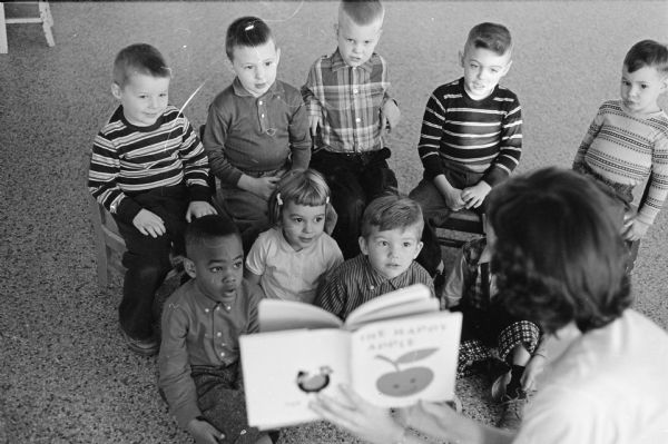 Story hour time at the Salvation Army all day nursery school, 121 West Wilson Street. Boys and girls are shown listening to a teacher reading from a book. Anne Shea served as the head teacher and was assisted by Mrs. Belva White. The school could accommodate up to twenty students ages 2 1/2 to 5 years old.
