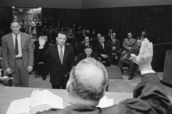 Dane County elected officials are swore into office for the 1961 term. District Attorney William D. Byrne (foreground) is shown with his hand raised to Dane County Circuit Court Judge Edwin M. Wilke swearing to conduct his duties in accordance with all laws.