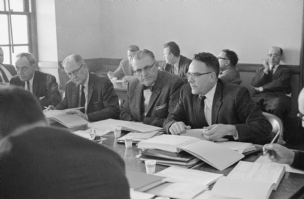 At left is Senator William Draheim, Neenah, chair of the Legislature's Joint Committee, and co-chair Everett Bidwell, Portage, center, listen to a budget explanation from Wallace Lemon, right, director of the Bureau of Management of the Department of Administration. (Others in the room are not identified.)
