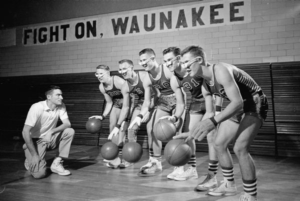 Head Coach Ron Hering (kneeling) poses with the starting five of the Waunakee High School basketball team. Members of the team shown (L-R) are Dave Roberts, Jerry Raemisch, Jerry Hackbart, Marv Hellenbrand and John Howard. The players practice a dribbling drill handicapped with goggles and gloves in preparation for their first-round game of the state WIAA district basketball tournament. The team has won all twenty of their games and is ranked #3 in the Little 16 ranking.