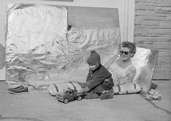 Mrs. Peter Berntsen, 926 LeRoy Road, enjoys an outdoor tanning session aided by a wall of aluminum foil. Her son Erik (3) plays in front of her bundled up in his winter coat. They were guests at the home of Mrs. F. J. (Mary) Davis at 424 New Castle Way. 
