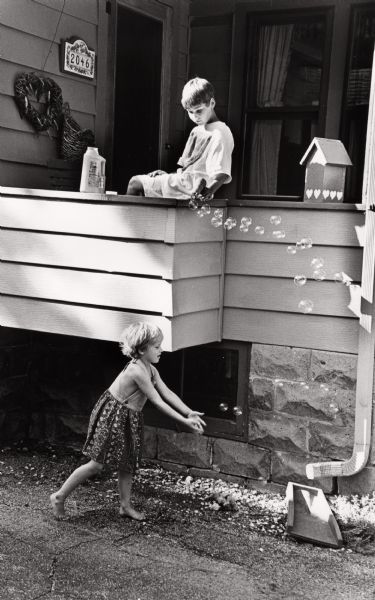 Two children playing with bubbles. A young boy sitting on the porch is making bubbles with a bubble gun, while a young girl on the sidewalk in the foreground tries to catch them.
