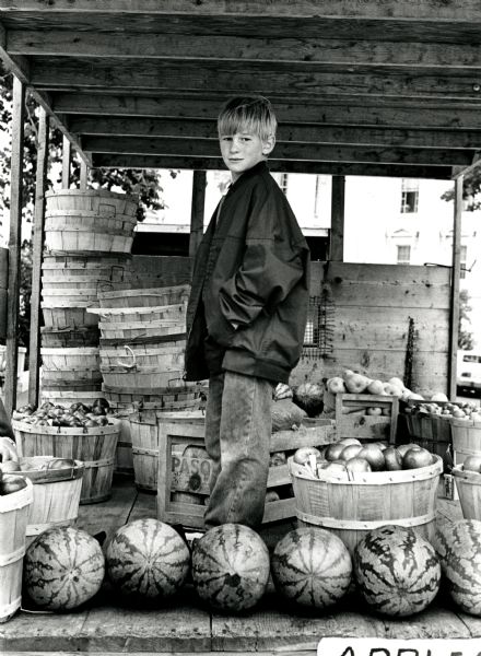 Boy posing with watermelons under a wooden stand at the Farmers' Market. The Wisconsin State Capitol is in the background.