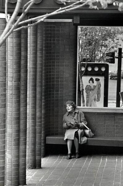 An elderly woman sitting on a bench while waiting for a bus in a bus shelter. Behind her is a window which looks down the street.