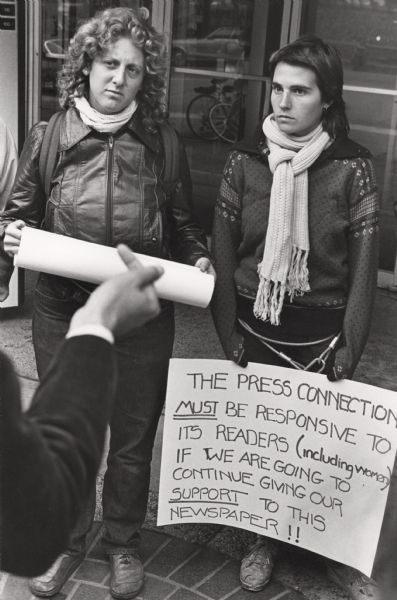 Lynette Margulies (left) and a friend holding signs and protesting against the "Madison Press Connection" newspaper. They are looking at a person standing in the left foreground.