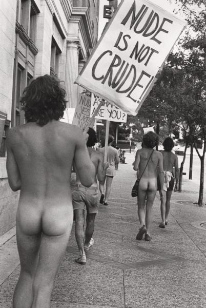 Rear view of several picketers walking nude down a sidewalk along State Street holding signs.