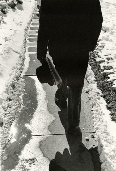 Rear view of a man, from the shoulders down, walking on a Doty Street sidewalk holding a hat and a newspaper in his hand. Snow is along both sides of the sidewalk, and the sun is reflected in melting snow and water.