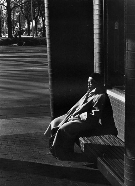 A woman sitting in a bus shelter while waiting for the bus at the Pinckney Street bus stop. Across the street people are walking along the sidewalk on Capitol Square.