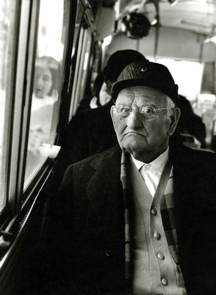 A man wearing eyeglasses and a hat travels on a bus.