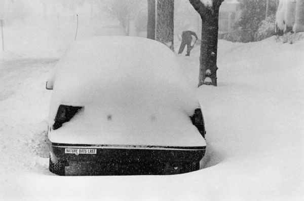 A car on Lakeland Avenue is buried in snow during a blizzard. A bumper sticker on the car reads: "Nature Bats Last." In the background a person is shoveling a sidewalk.