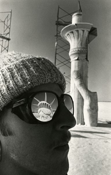 Close-up of a man gazing at the Statue of Liberty installation on a frozen and snow-covered Lake Mendota. The sculpture was constructed by the Pail and Shovel party of the University of Wisconsin. In the background is the hand and torch section of the sculpture near scaffolding. The head of the statue is reflected in the lens of the man's sunglasses.