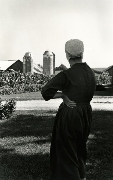 Amish woman looking at farm buildings and two silos.
