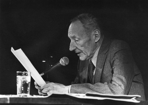 Writer William S. Burroughs seated and speaking into a microphone during a reading.