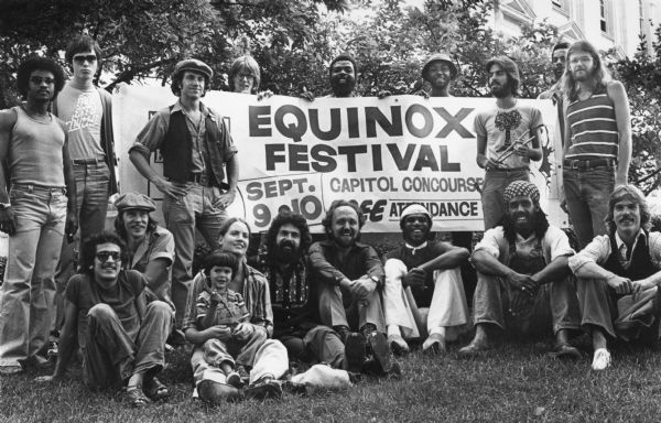A group portrait of musicians posing with a banner at the Equinox Festival. Seated in front from left to right are Tony Castaneda, drummer Ken Koepler (kneeling by Castaneda), an unidentified woman and child, Marcos Gonzalez, Rob Corbit, Merrit Mapp, Tony Brown, and drummer Jim (Pie) Cowan. Standing in back from left to right are an unidentified man, piano player Pat Wick, Gage Averil, Chaz Moore, two unidentified men, drummer Arno Gonzalez, bassist Paul Backstrom, and, behind Backstrom, bassist Ted Wingfield. Marcos Gonzalez, Corbit, Wingfield, and Brown played in the Tony Brown Band. Wick, Arno Gonzalez, and Castaneda played in Olmeca. Averil and Moore played in Irish Brigade. Moore was the organizer of the festival. 