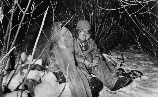A homeless man in Milwaukee sitting in the snow as he attempts to craft a makeshift shelter from a piece of tarp.