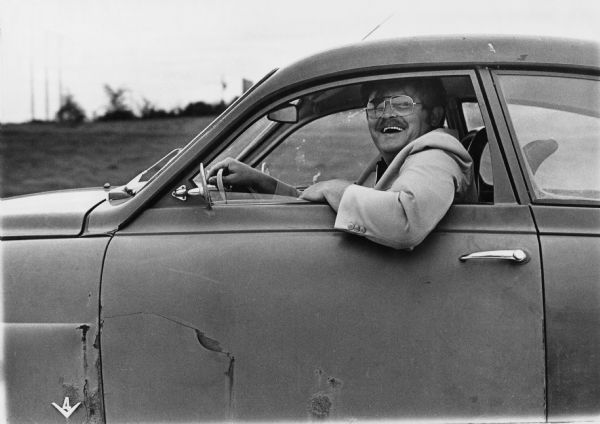 A smiling man driving I-90 in his rusty car.