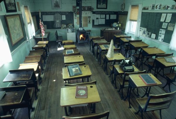 Back of a classroom at the Old Halfway Prairie Schoolhouse, a one-room schoolhouse, which operated from 1844 until 1961. In the front of the room is a woodburning stove. Set on some of the desks are books, a dunce cap, a typewriter, a slate, and a chalkboard with an abacus.