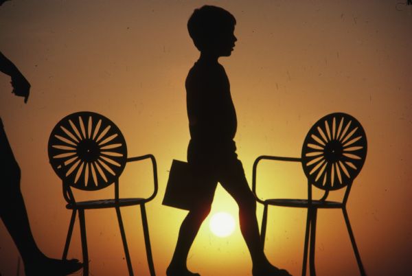 Silhouette against the setting sun of a young boy walking along the Memorial Union Terrace between two chairs.