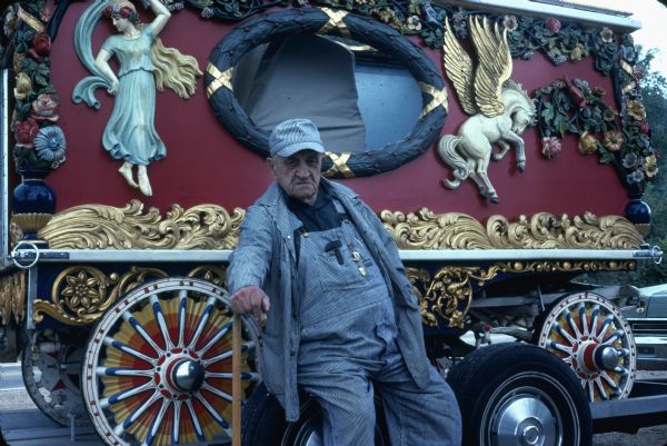 A man poses in front of a calliope, a steam-powered pipe instrument in a large, ornately decorated circus wagon.