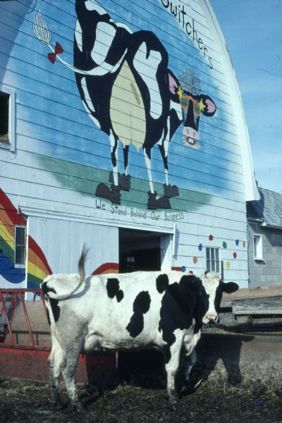 A dairy cow in front of a large mural on the side of a barn. Below the mural is written: "We Stand Behind Our Business."