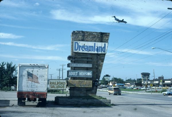 Truck stop sign that reads: "Dreamland" which is north of Milwaukee. Across the street is the Wauwatosa Savings and the Tri City National Bank. An airplane is flying overhead.