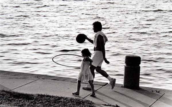 View looking down at a father and daughter walking on the sidewalk along the shore of Lake Mendota at James Madison Park. The father is playing with a basketball, and the young girl is playing with a hula hoop.