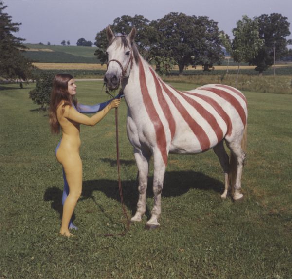 A nude female model painted partially with blue paint is standing outdoors holding the bridle of a horse painted with red and white stripes. The reverse side of the horse (unseen) is painted with polka dots. The woman was referred to as the "Lady on the Horse."