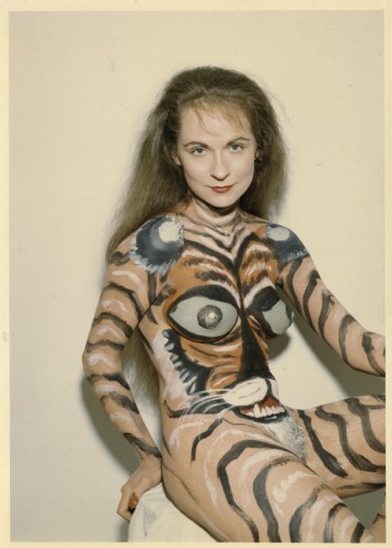 Three-quarter length portrait of "Tiger Woman." Sitting on a stool before a white wall, the long-haired model states straight at Sid, the photographer. 