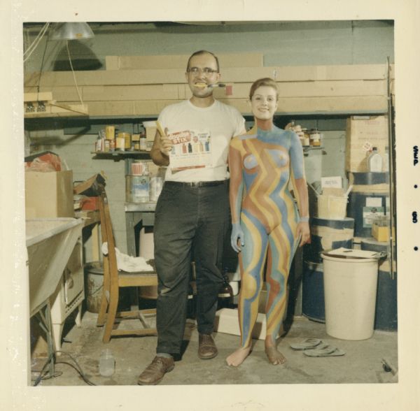 Gene Coffman, with a paintbrush in his mouth and holding a package of  Disguise Stix©, standing next to "Abstract Girl" in his garage on Sommers Avenue. The pattern, painted by Sid on the model's body, was inspired by an original work by Sid titled: "Traffic."