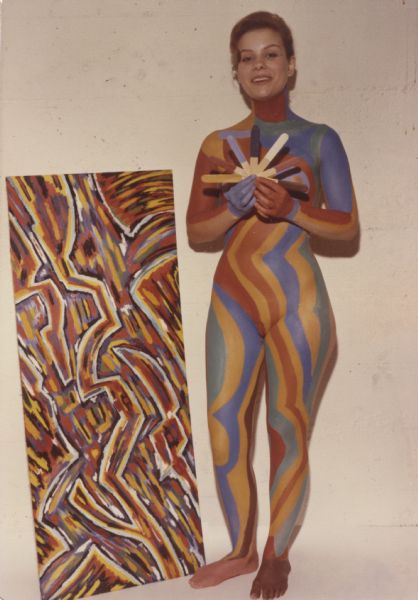Full-length portrait of "Abstract Girl" standing next to an abstract painting titled "Traffic" by artist, Sid Boyum. The painting was used as inspiration for the pattern on the model. She is holding a representative group of Disguise Stix©, the body paint developed by Gene Coffman, of Gratco Corporation.