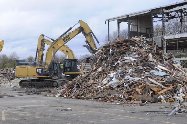 View from the north side, looking east, of the State of Wisconsin's Central Services Building, located at 201 S. Dickinson Street. The building contained Transportation Services and the UW Tandem Press. It is being torn down in preparation for building a new storage facility, the State Archives Preservation Facility (SAPF). In the foreground is a large mound of debris, and on the left is a man standing on one of two Caterpillar 349E Hydraulic Excavators.