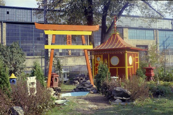 View of Sid Boyum's backyard, with an arch, pagoda, and sculptures among shrubs and trees. There is an empty pond under the arch. The Madison-Kipp Corporation factory is in the background. 